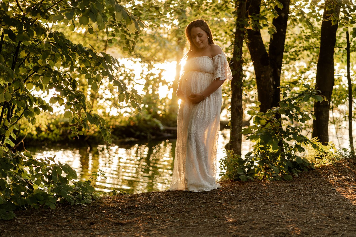 Pregnant woman in white dress standing among trees near sunlit lake at dusk.