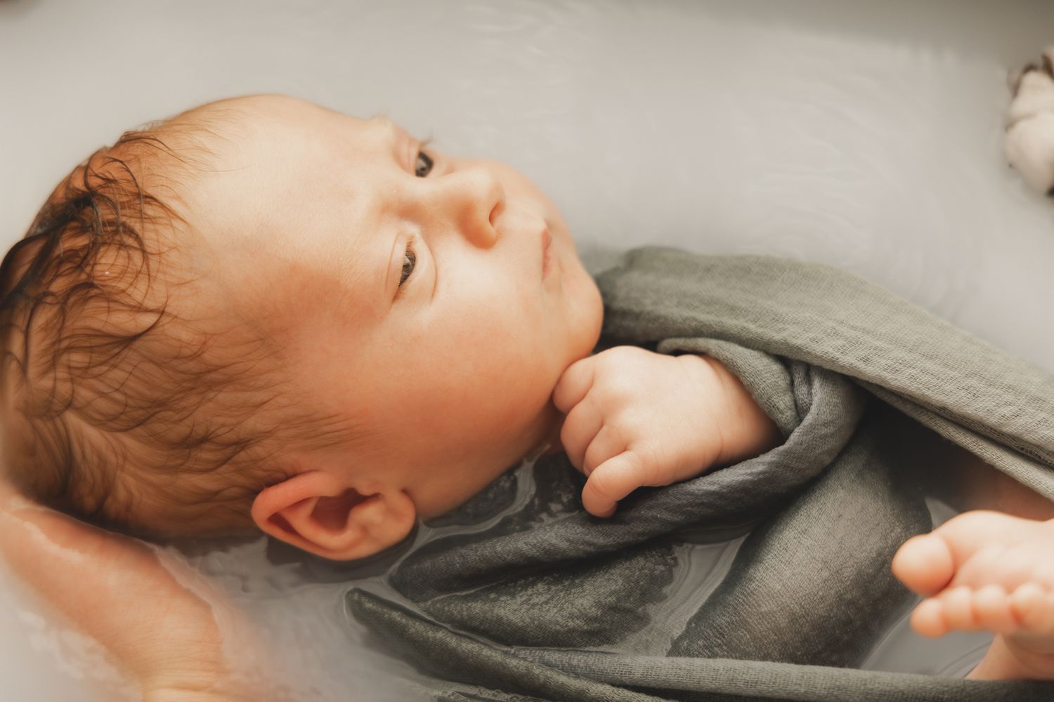 Séance photo nouveau-né en Isère : l'apaisement d'un bébé emmailloté dans un lange gris lors d'un bain de lait.