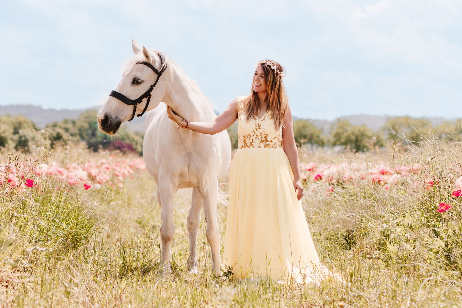 Woman in yellow dress walking with white horse through colorful wildflower meadow.