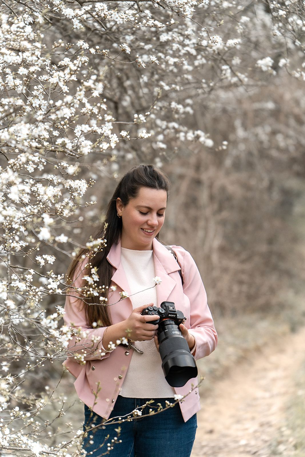 Woman in pink jacket reviewing photos on camera beside white flowering spring trees.