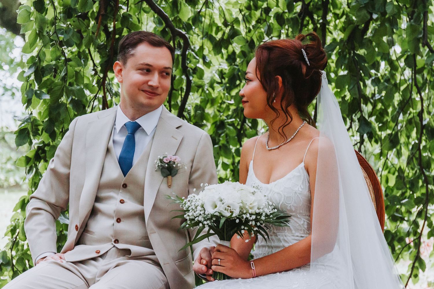 Newlywed couple sitting together under green arch, groom in tan suit, bride holding white bouquet.