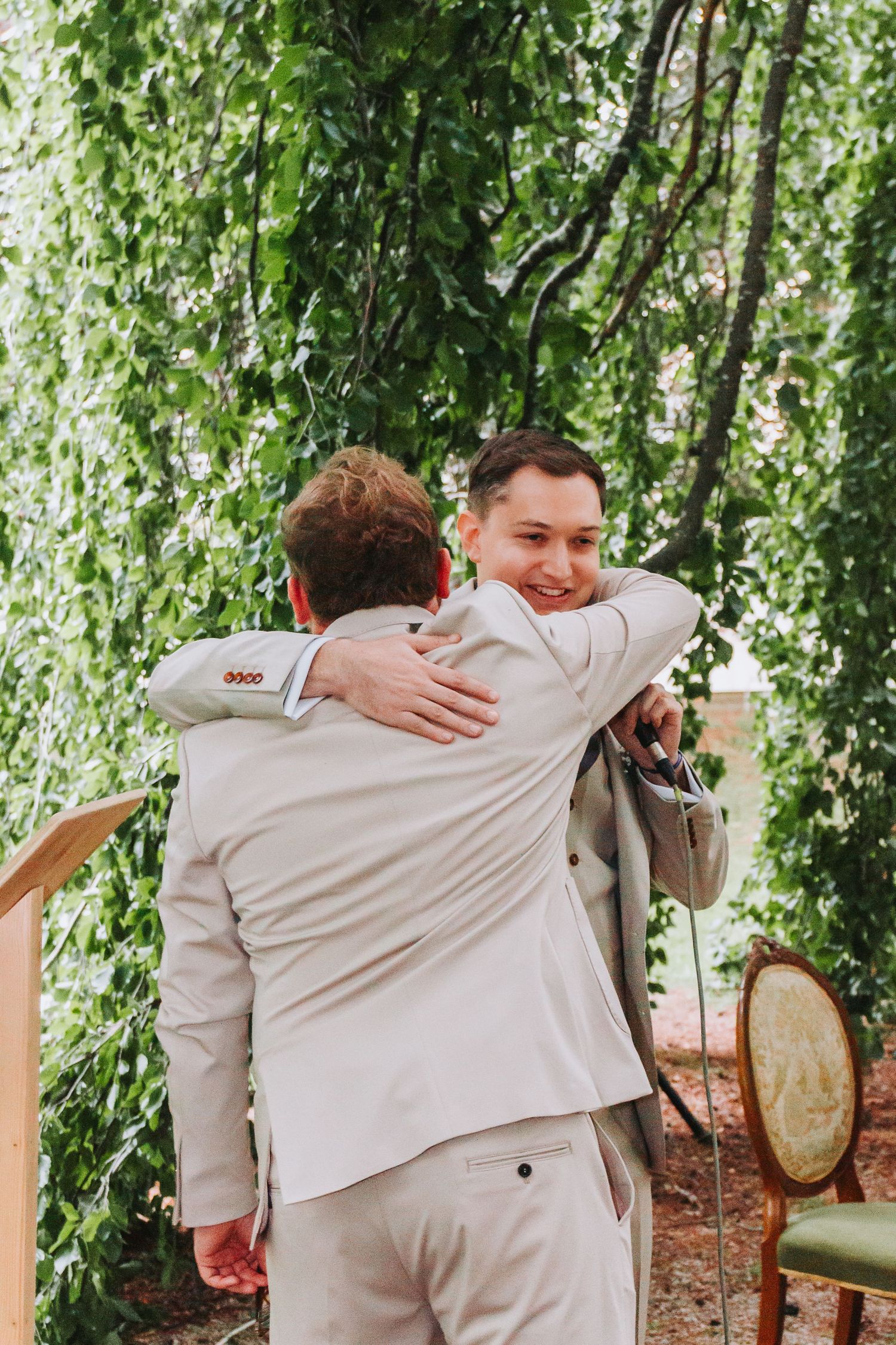 Two groomsmen in beige suits share a warm embrace during an outdoor wedding ceremony under green hanging branches.