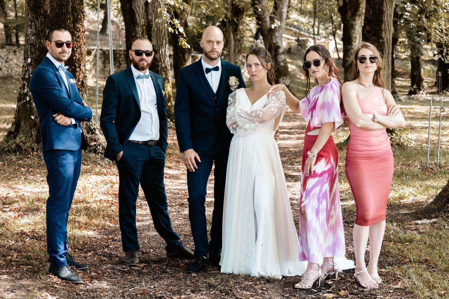 Wedding party of six posing casually in formal attire among trees at outdoor venue.