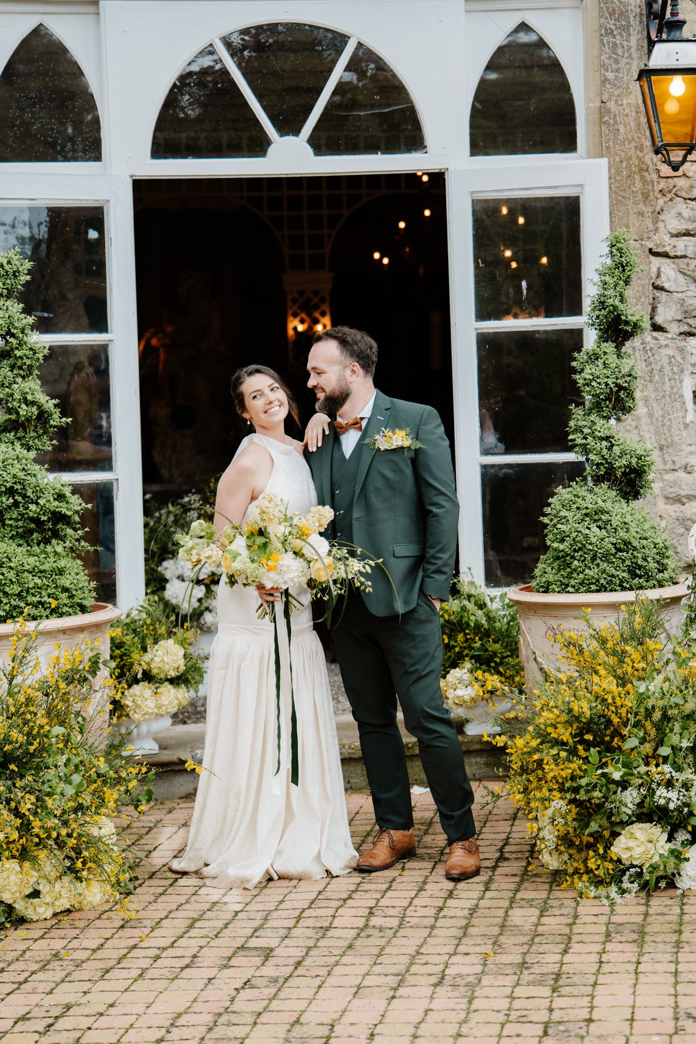 Bride and groom posing together outside a venue, bride holding yellow and white bouquet in wedding dress.