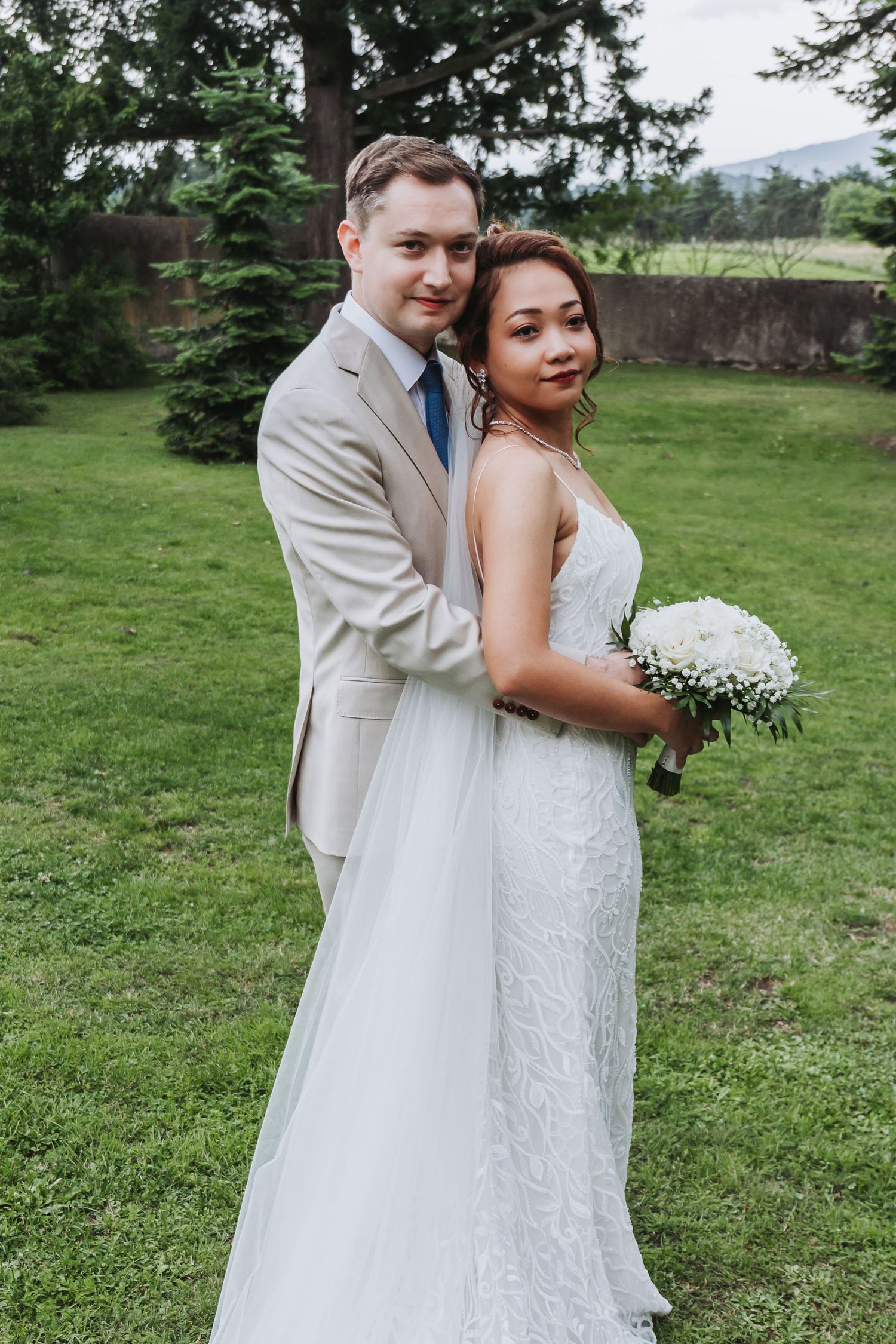 Interracial wedding couple embracing on green lawn, bride in lace gown holding white bouquet, groom in beige suit with blue tie.
