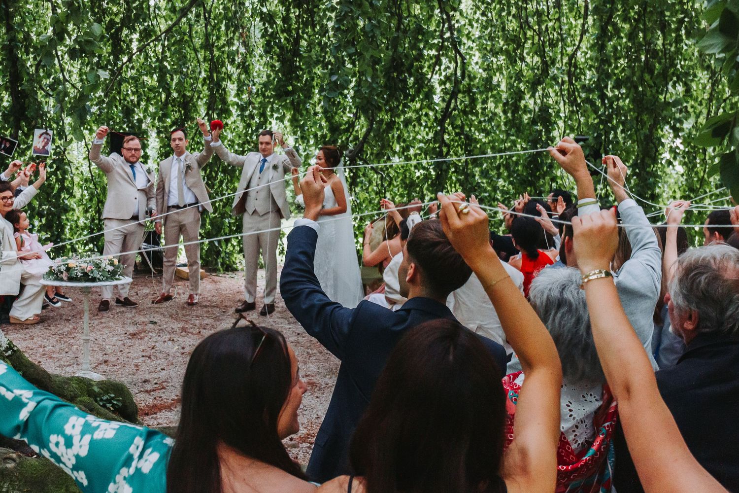 Wedding guests raise hands in celebration during outdoor ceremony beneath cascading green willow branches.