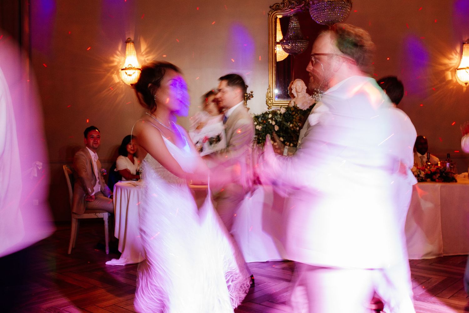Bride dancing energetically with guests under purple lights at lively wedding reception.