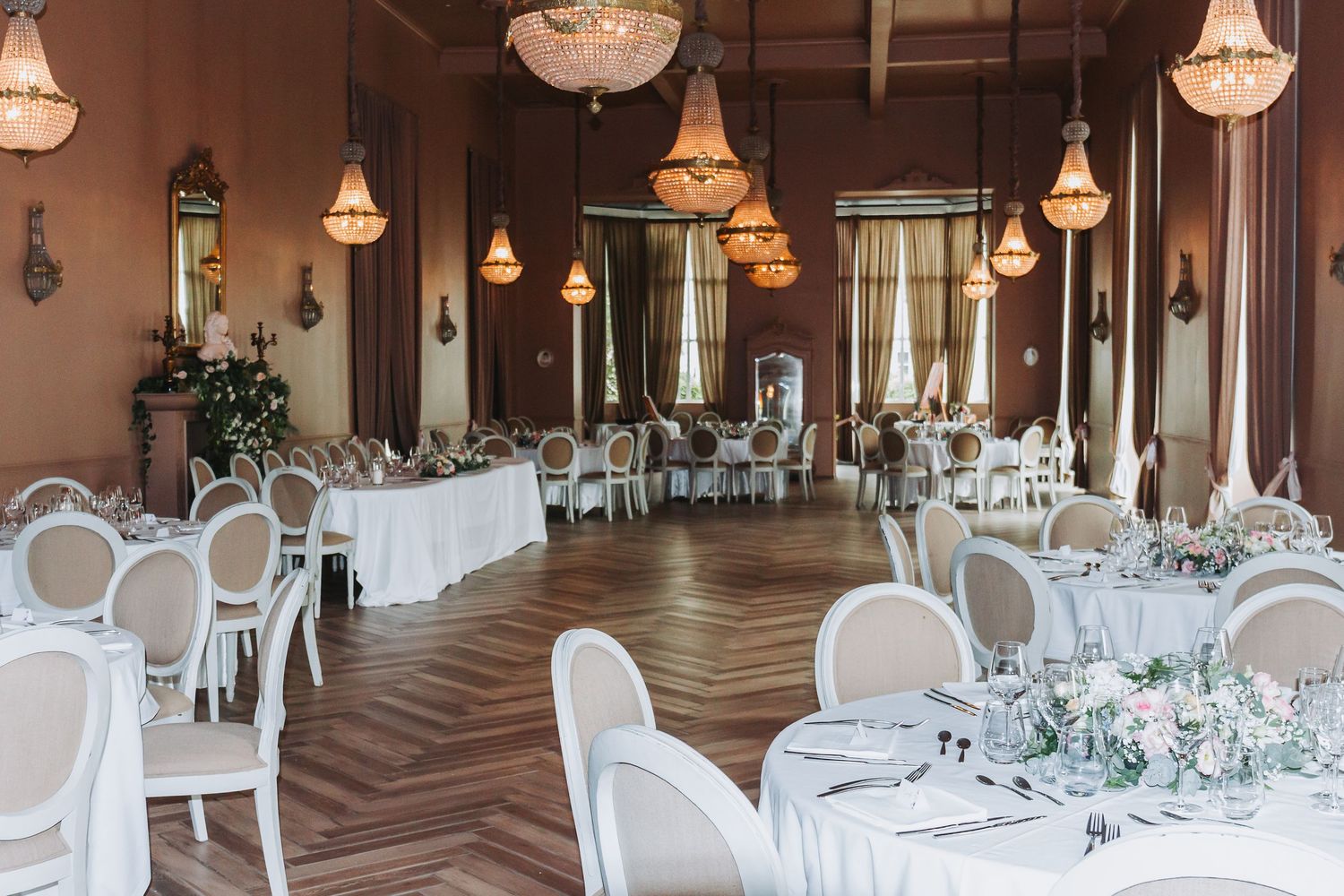 Grand wedding reception ballroom with crystal chandeliers, round tables, and herringbone wood floors.