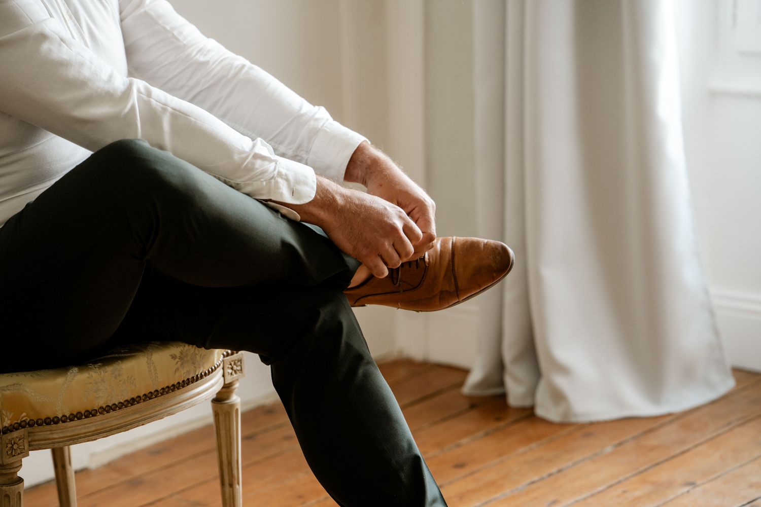 Groom sitting on a chair putting on brown leather dress shoes while wearing dark trousers and white shirt.