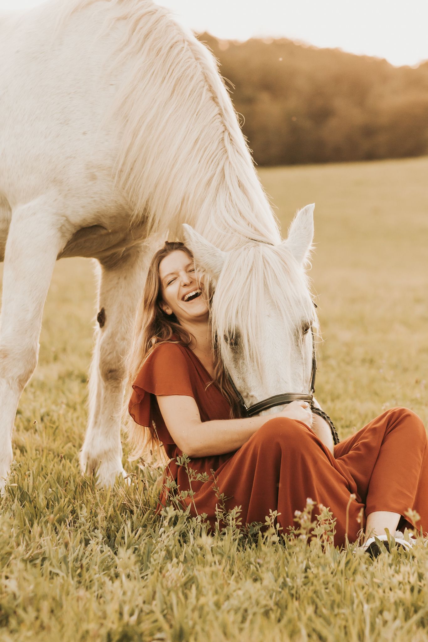 A laughing woman in red sits beside a grazing white horse in golden light.
