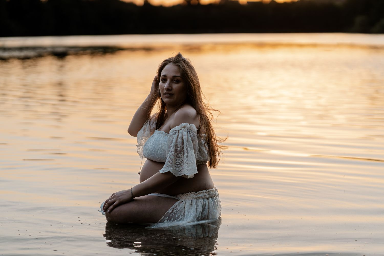 Pregnant woman in white lace ensemble sitting serenely in shallow lake water at golden hour sunset.