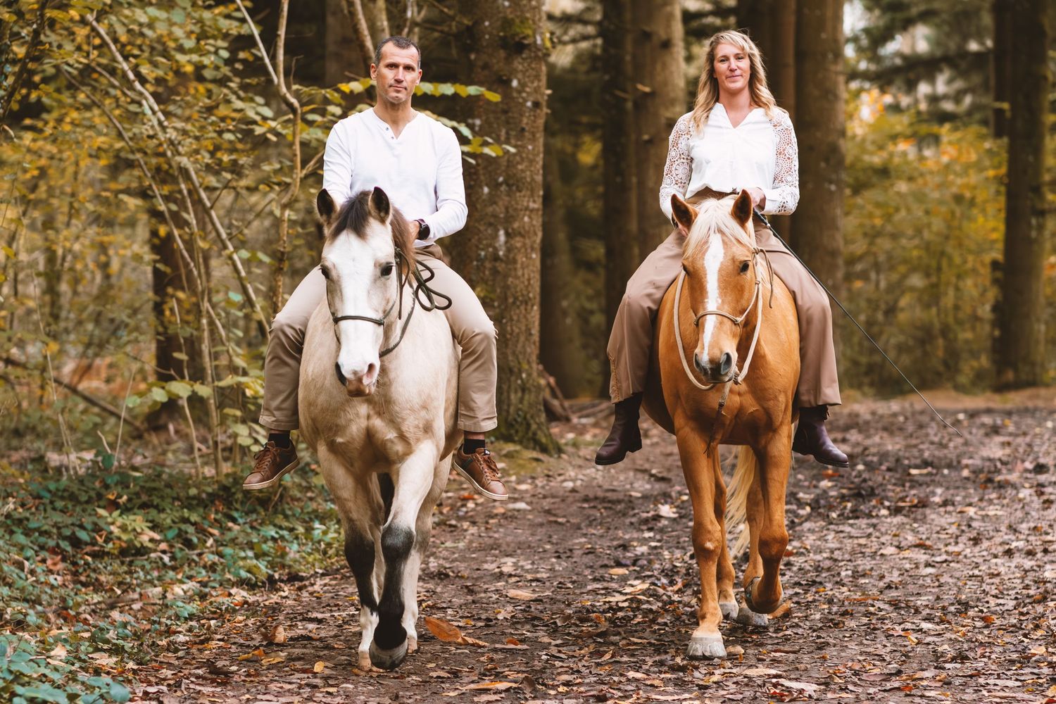 Couple in white shirts riding two horses toward camera on a leaf-covered forest trail.