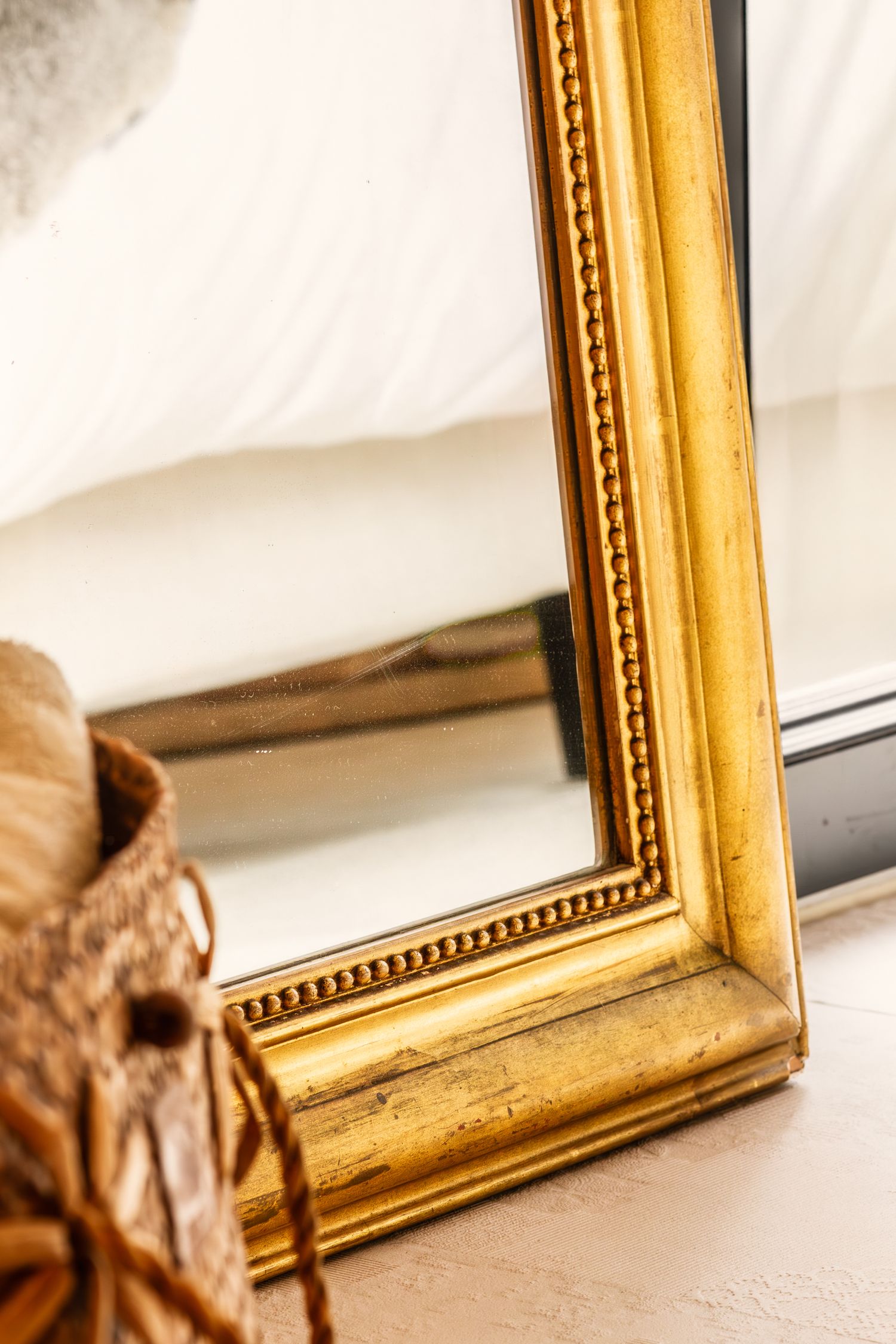 Close-up of a decorative gold-framed mirror leaning against a white wall beside a wicker basket.