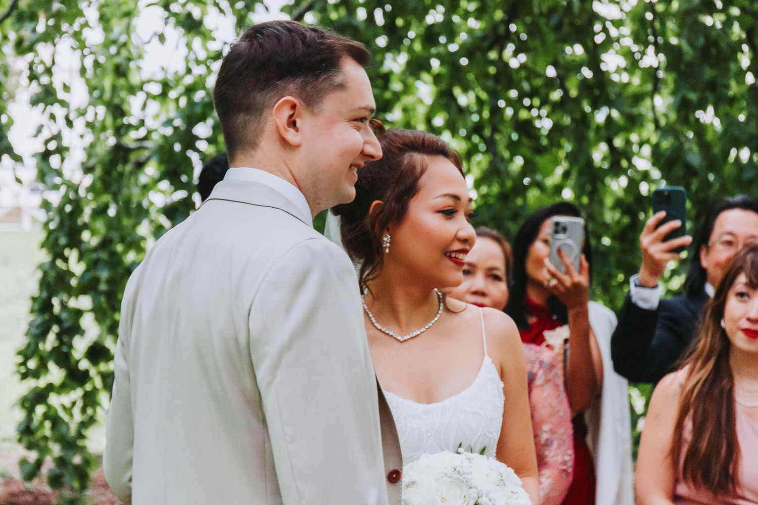 Smiling newlywed couple during outdoor wedding ceremony surrounded by lush green foliage and guests.