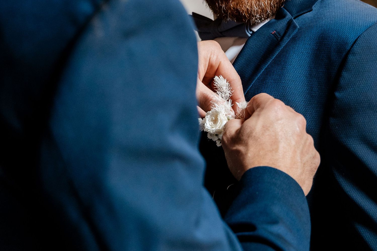 Groomsman pinning a white boutonniere onto a groom's navy blue suit lapel at a wedding.