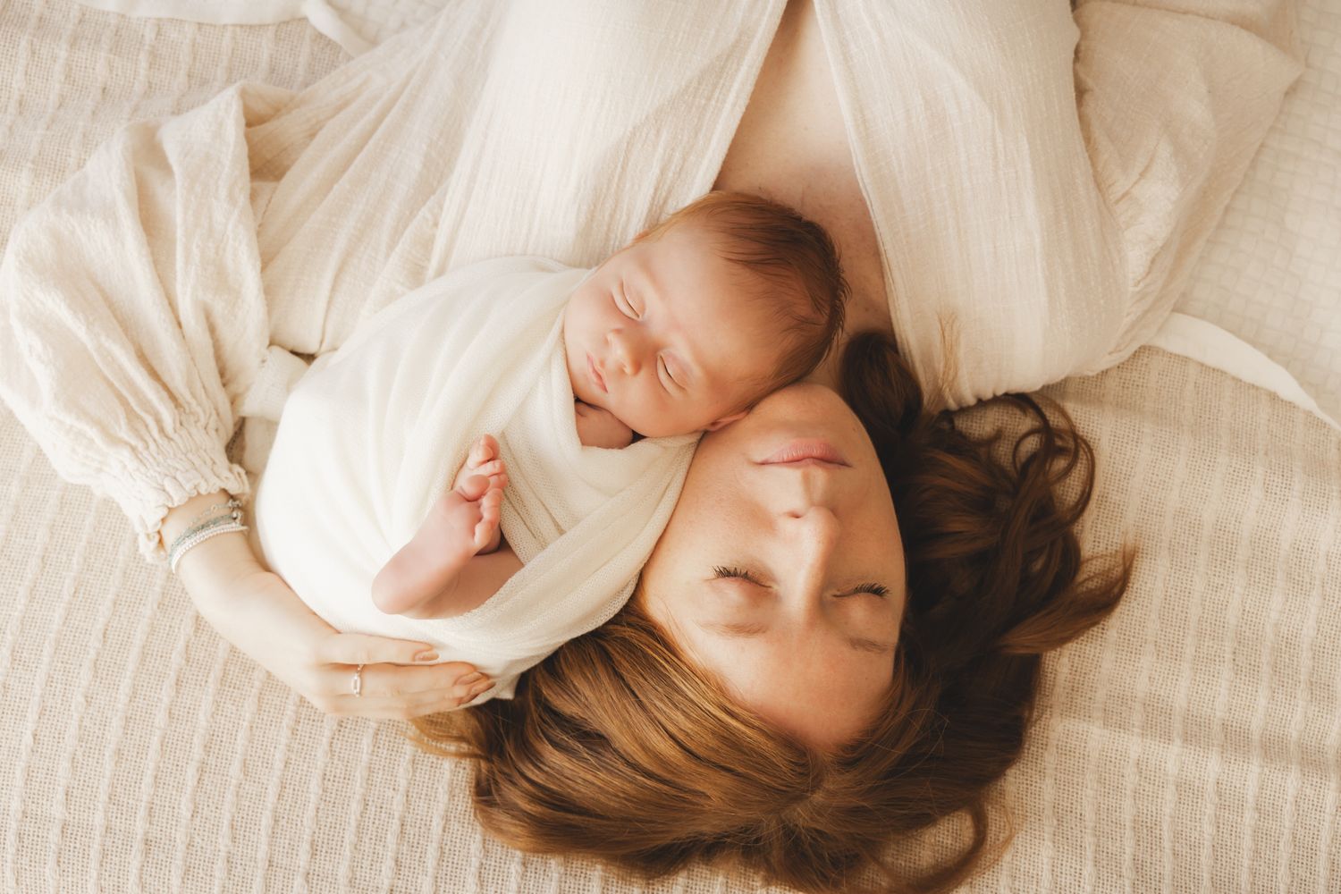 Mother lying down with eyes closed, tenderly holding sleeping newborn baby wrapped in white swaddle.