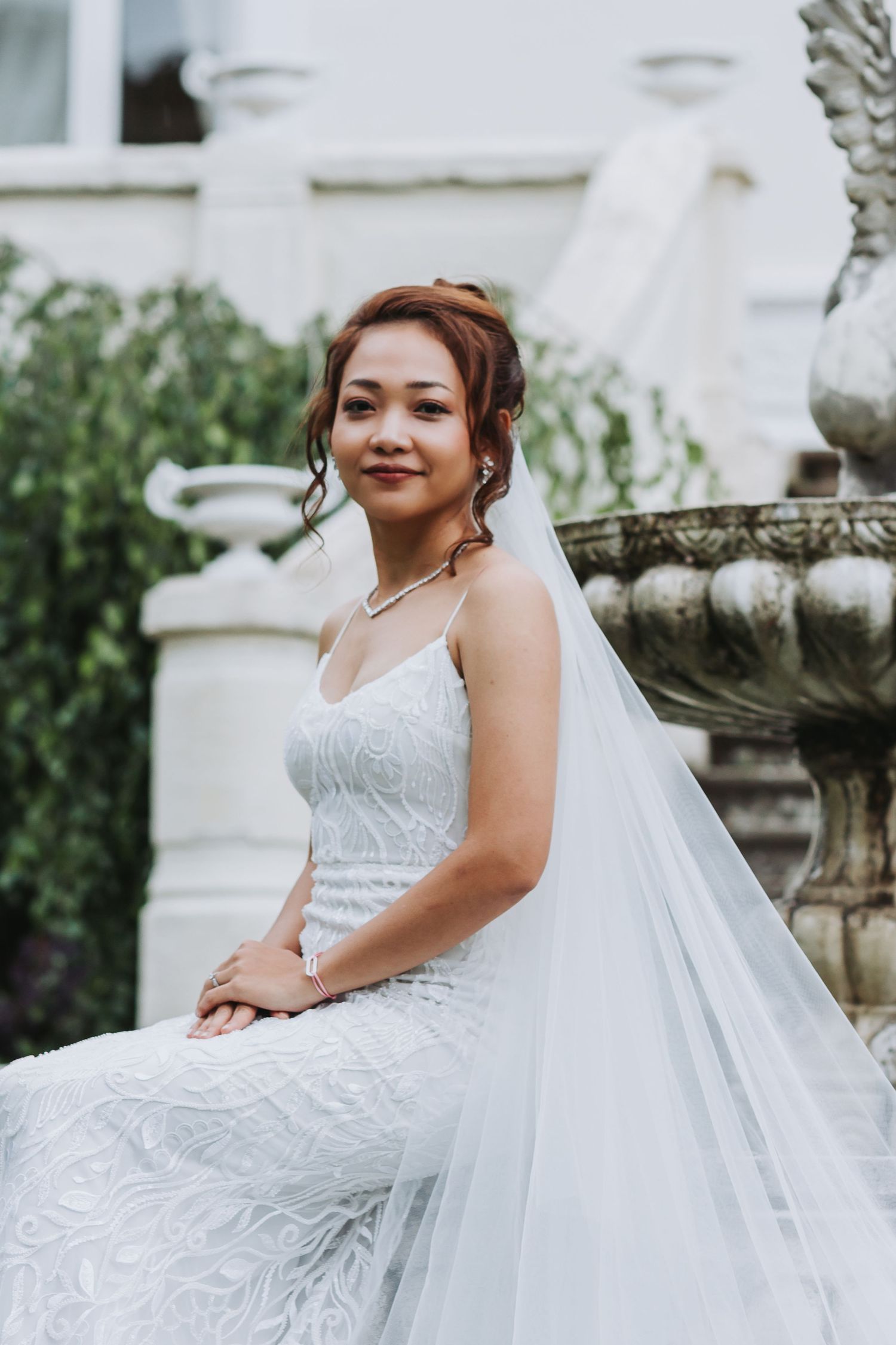 A bride in a white lace gown and veil sits gracefully outdoors near stone pillars, smiling softly.