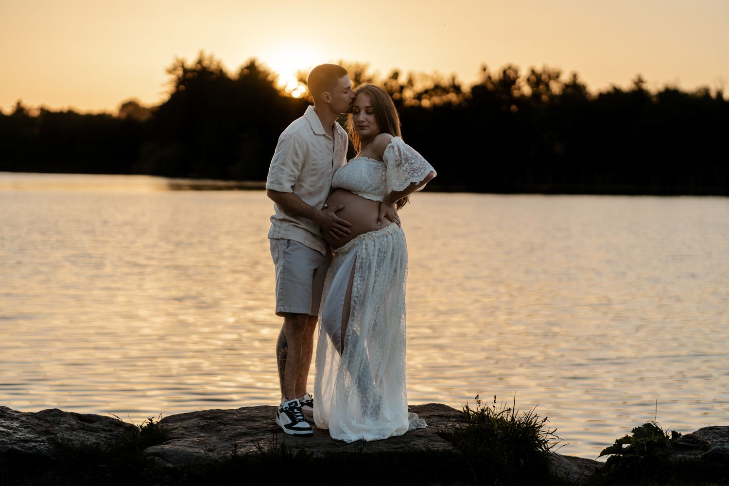 Man embracing pregnant woman in white dress on rocks by lake at sunset.