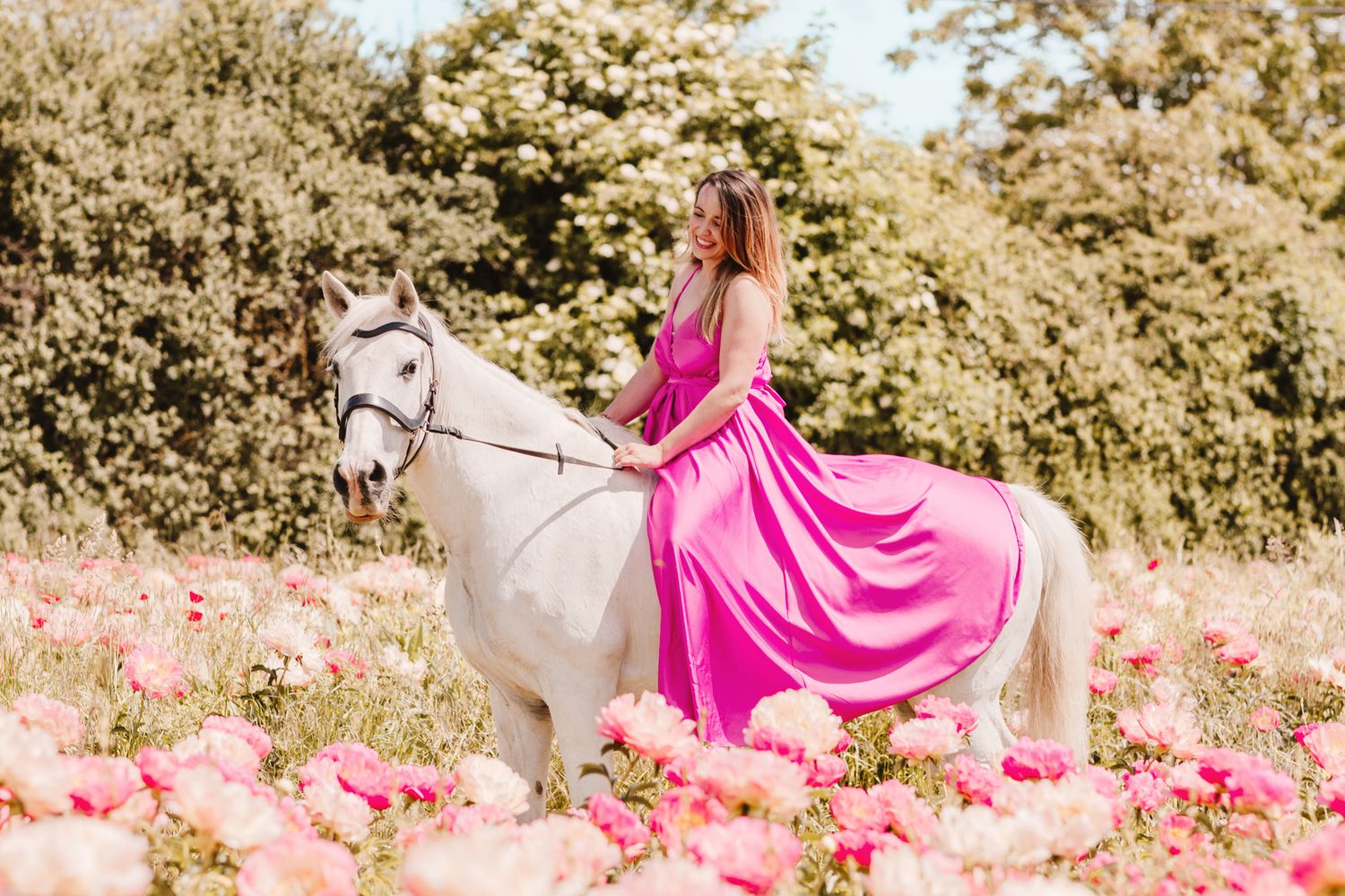 Woman in pink dress riding white horse through blooming pink flower field.