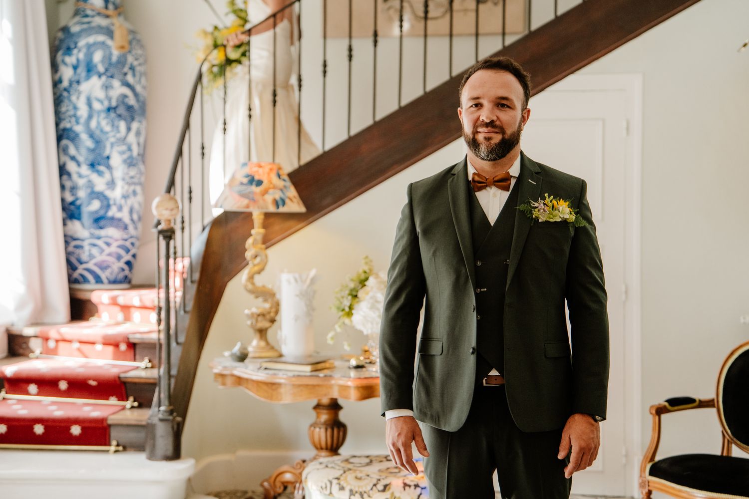 Smiling groom in dark green three-piece suit with floral boutonniere standing near a staircase indoors.
