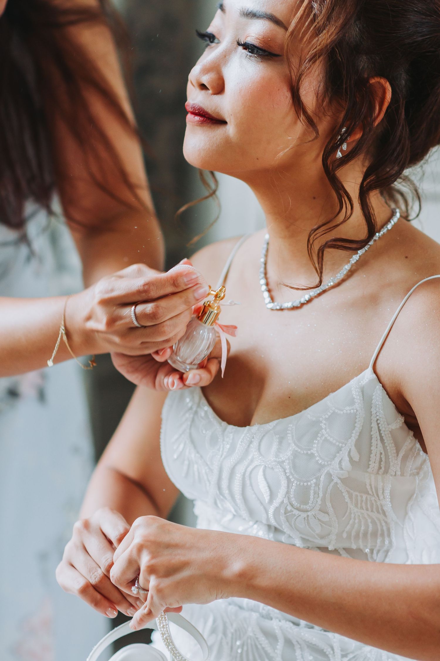 Asian bride getting perfume applied by bridesmaid while wearing a white lace dress and pearl necklace.