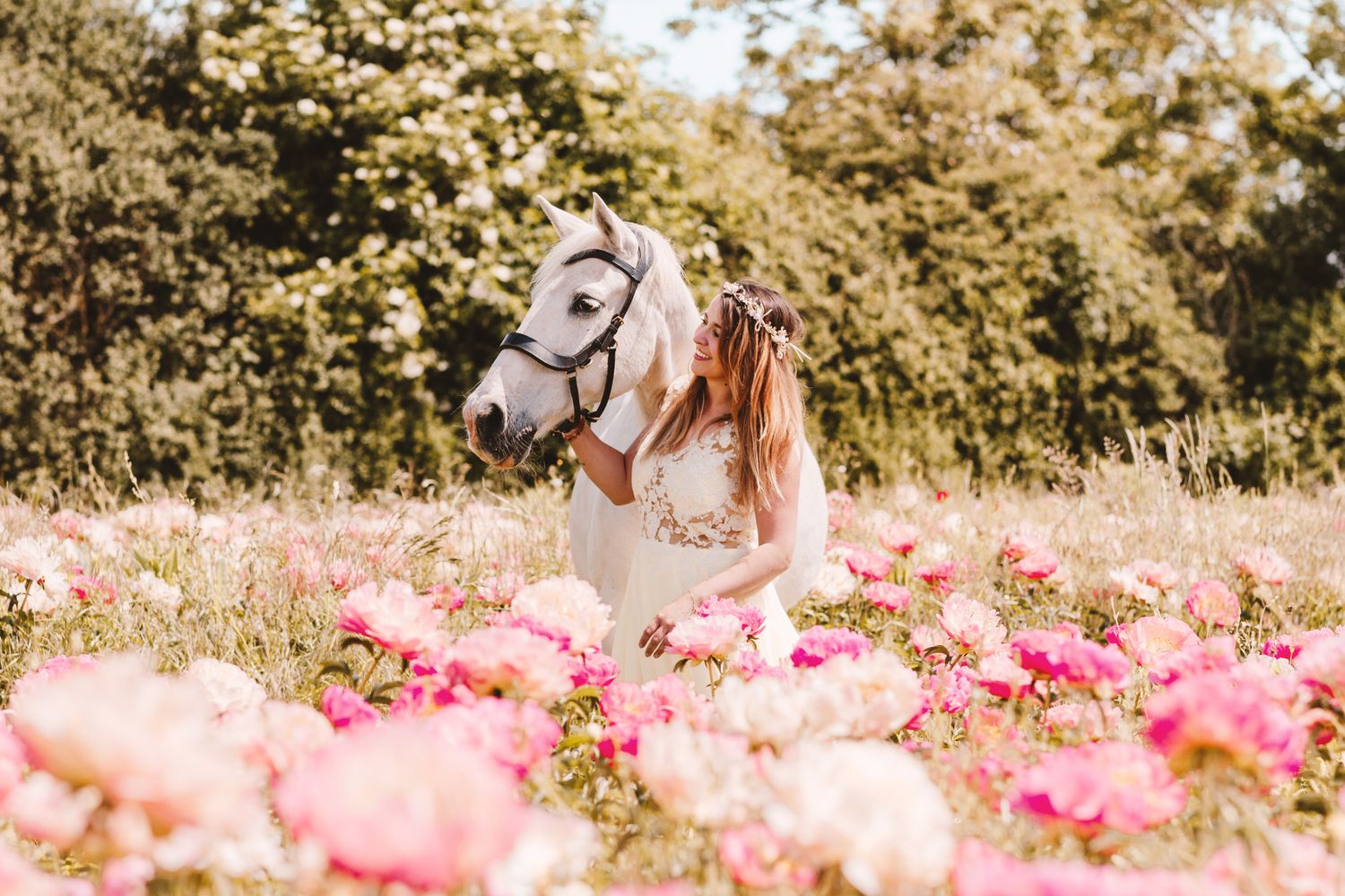 Woman in white dress embracing white horse surrounded by pink peonies in field.