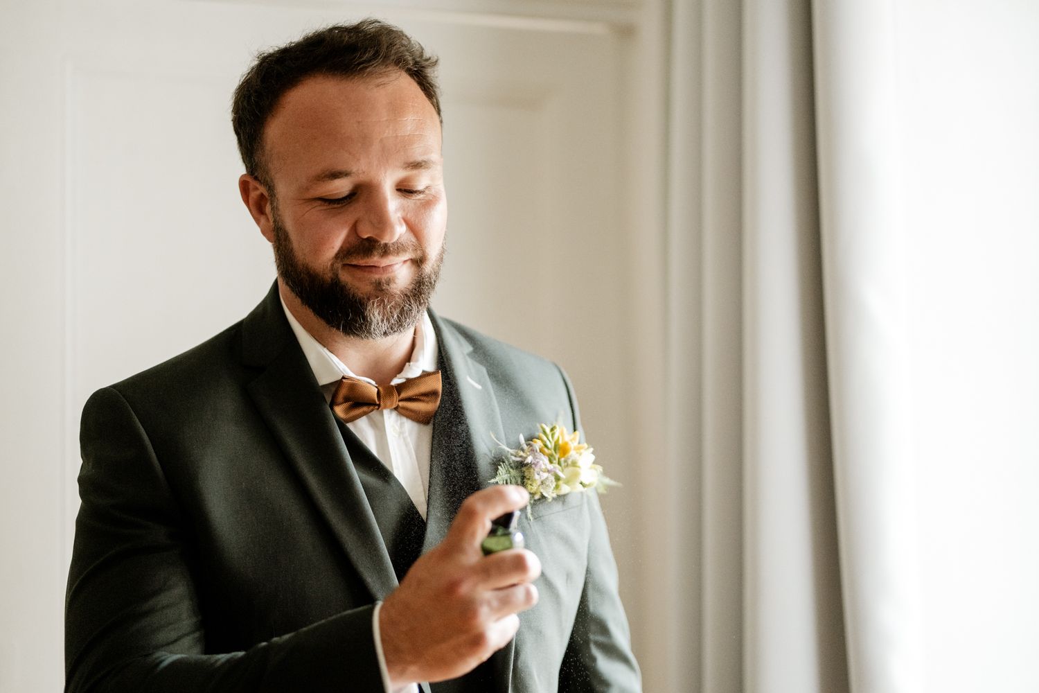 Bearded groom in dark green suit with wooden bow tie adjusting white daisy boutonniere.