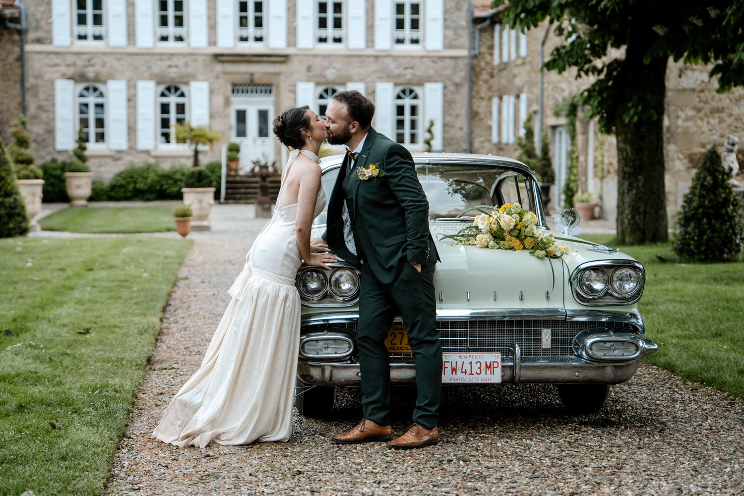 Bride and groom kissing beside a vintage teal Cadillac in front of a French chateau with floral decorations.