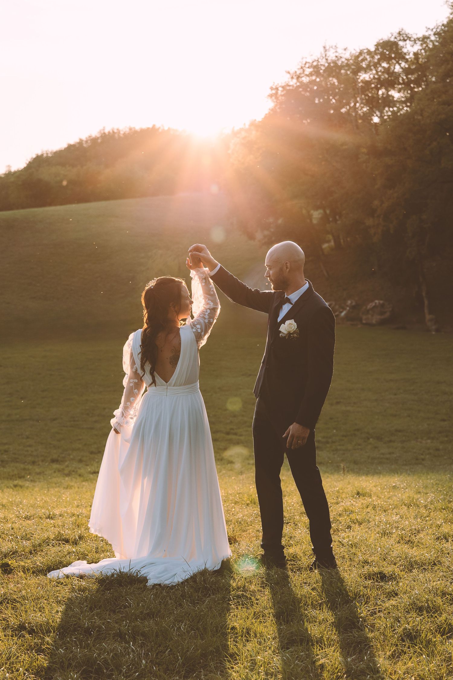 Groom twirls bride in flowing white gown on green meadow during stunning golden sunset wedding portrait.