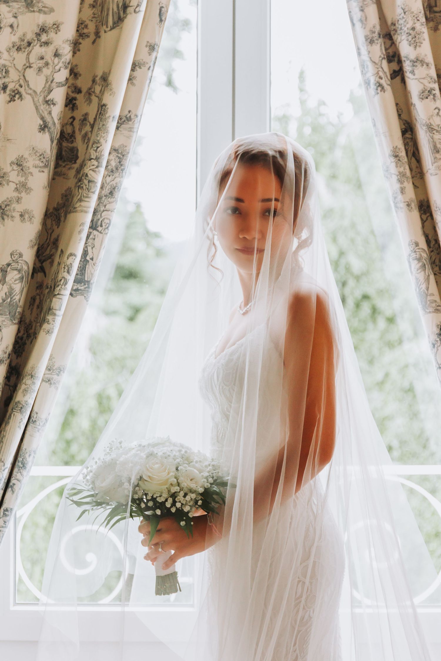 Bride veiled in white tulle holding a white bouquet standing by a window with floral curtains.