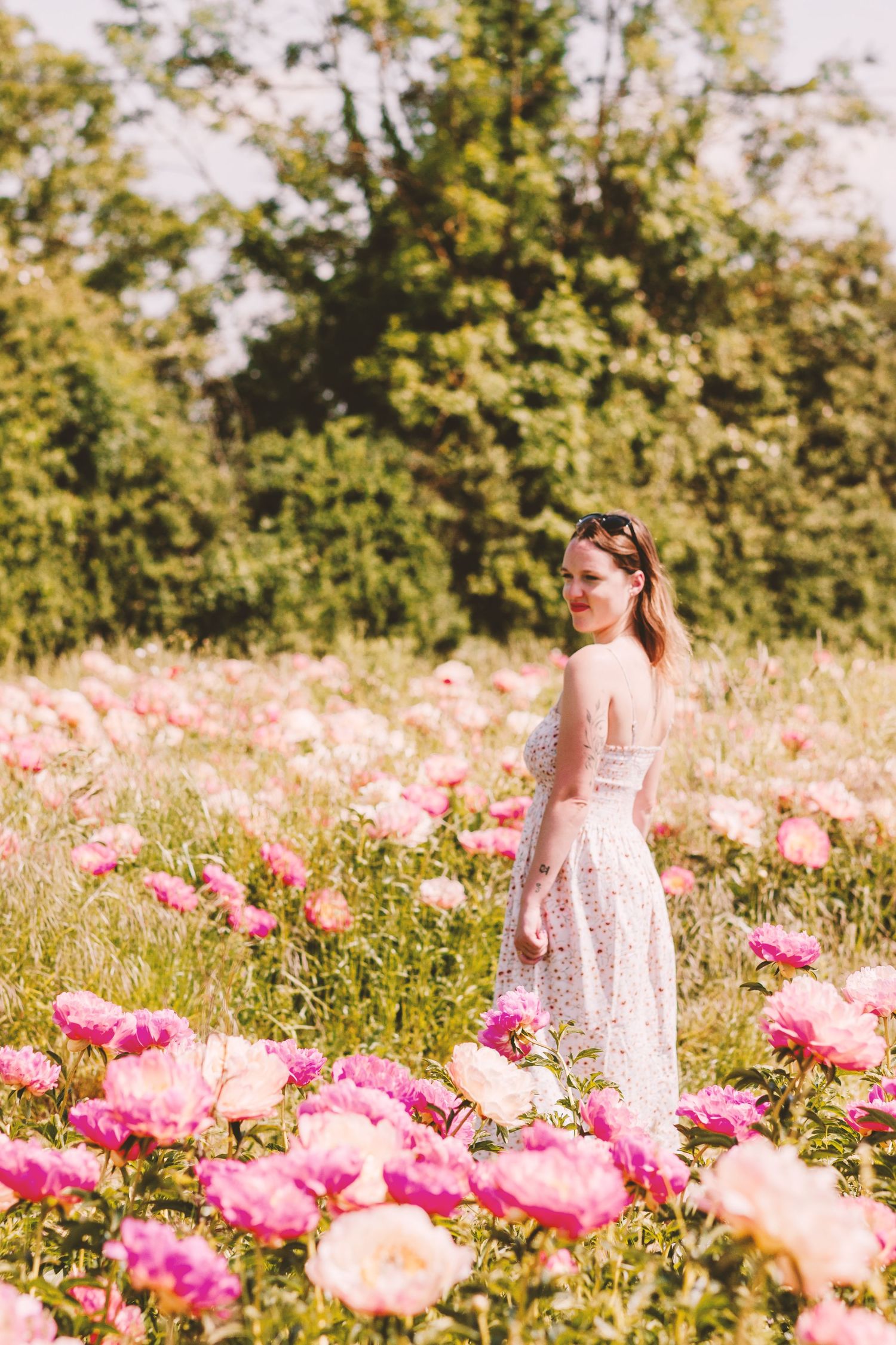Young woman in white dress standing among vibrant pink peonies in summer field.