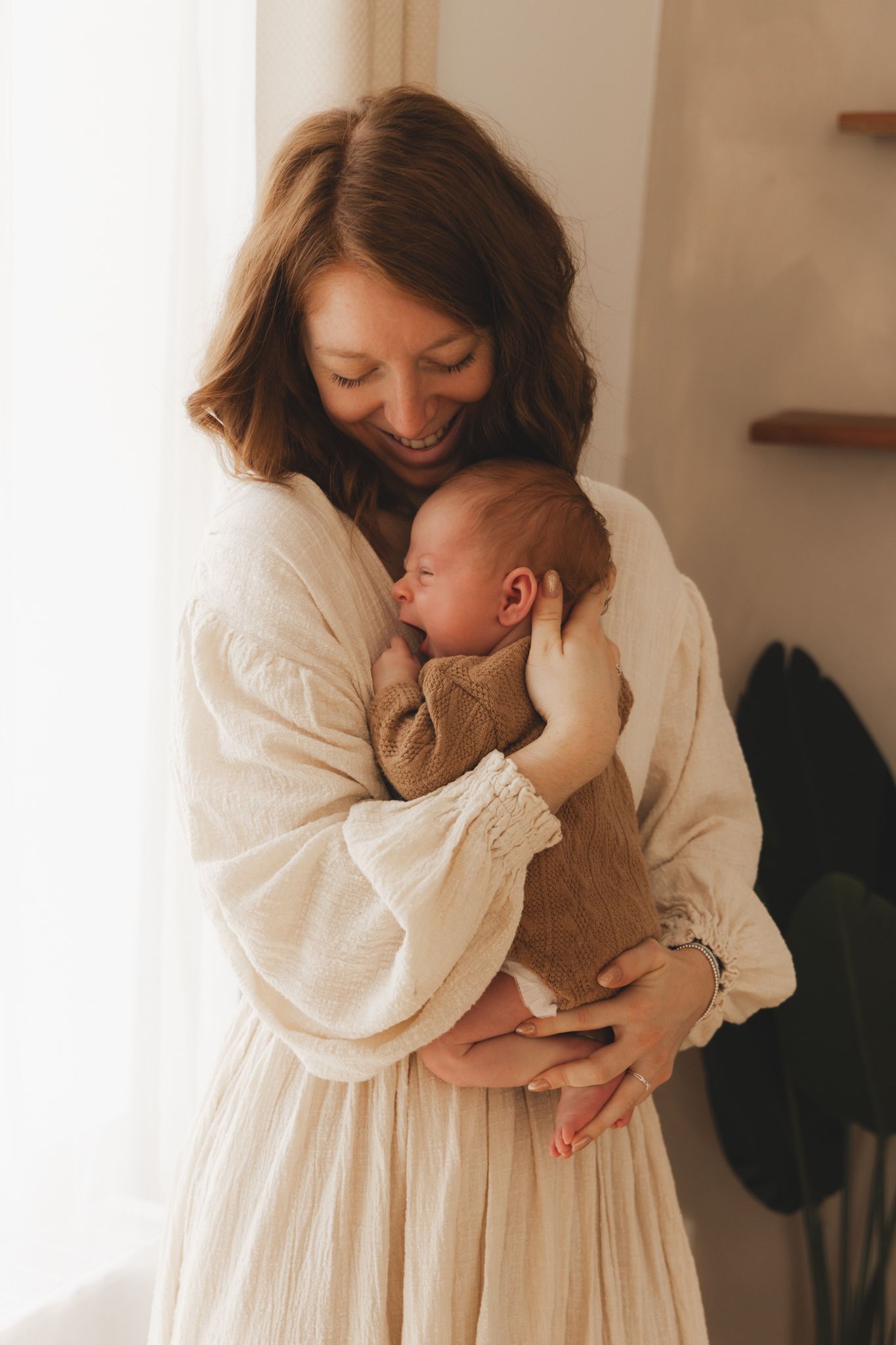 Smiling mother in white linen dress tenderly holding sleeping newborn baby near a bright window.