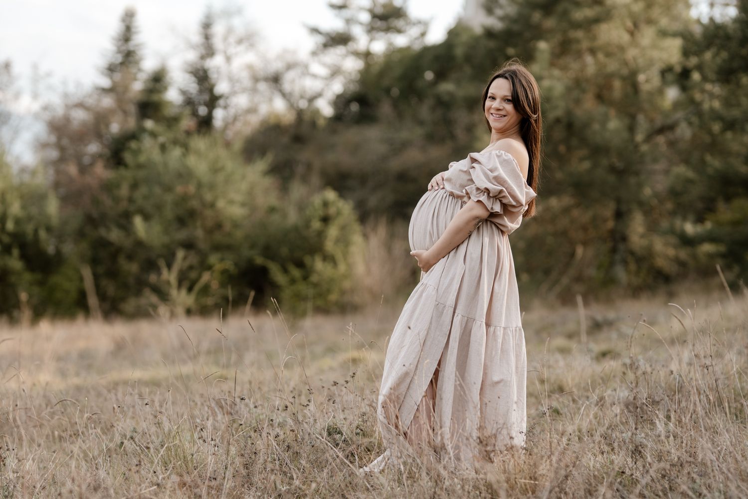 Photo de grossesse en Isère : le sourire lumineux d'une future maman en robe beige au cœur d'une prairie aux couleurs d'automne.