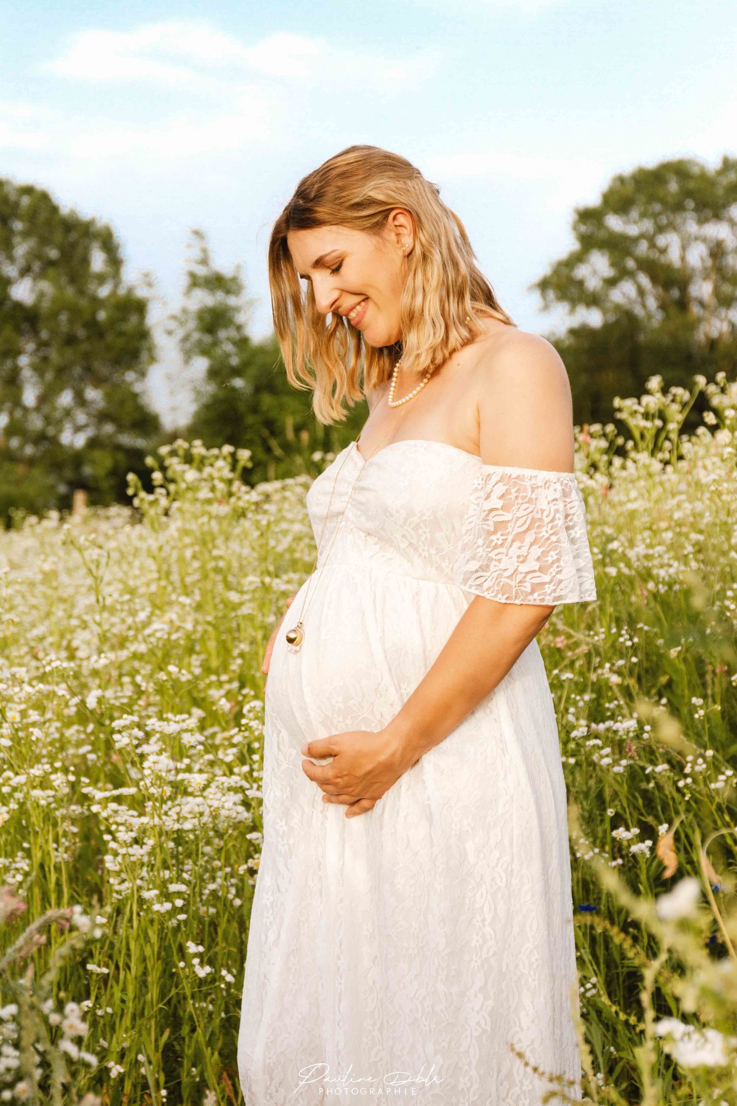 Smiling pregnant blonde woman in white lace dress cradling her bump in a wildflower meadow.
