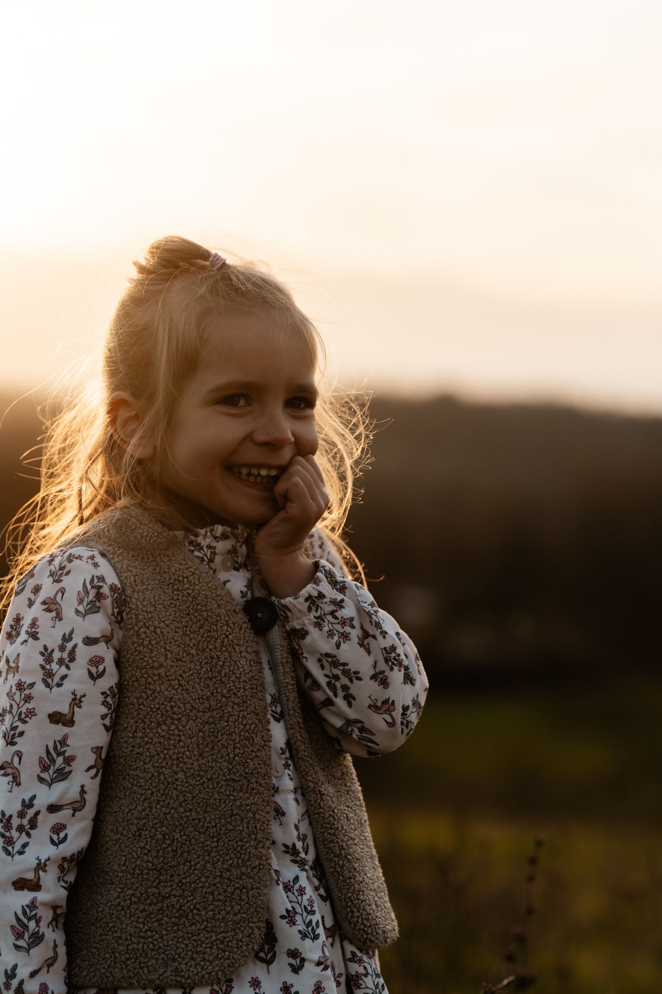 Séance photo enfant en Isère : une adorable petite fille blonde en robe à fleurs sourit au coucher du soleil.