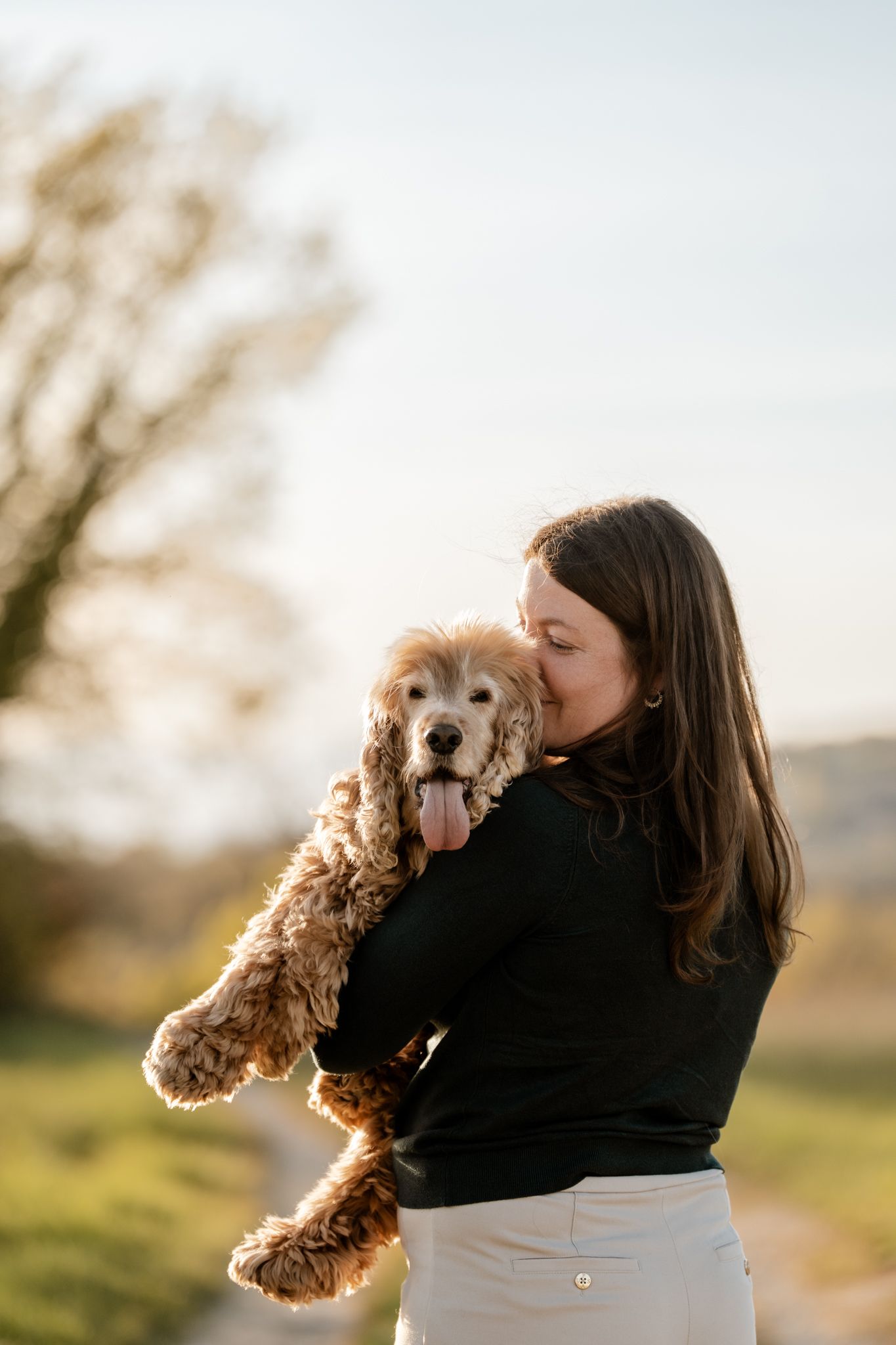 A dark-haired woman in a black sweater cuddles a fluffy Cocker Spaniel outdoors at golden hour.