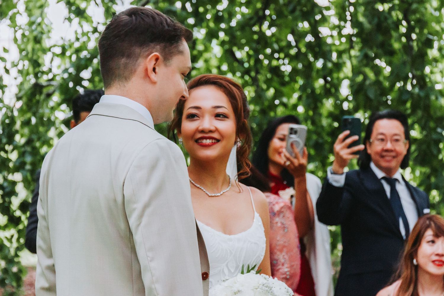 Happy bride smiling at groom during outdoor wedding ceremony with green leafy backdrop and guests.