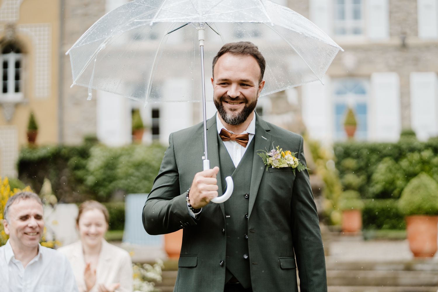 Happy groom in green three-piece suit holding transparent umbrella at elegant outdoor wedding venue.