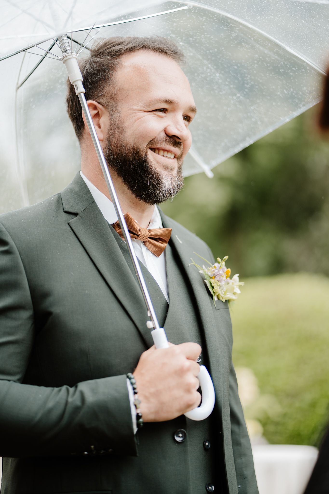 Smiling bearded groom in green suit holding a clear umbrella outdoors with a floral boutonniere.