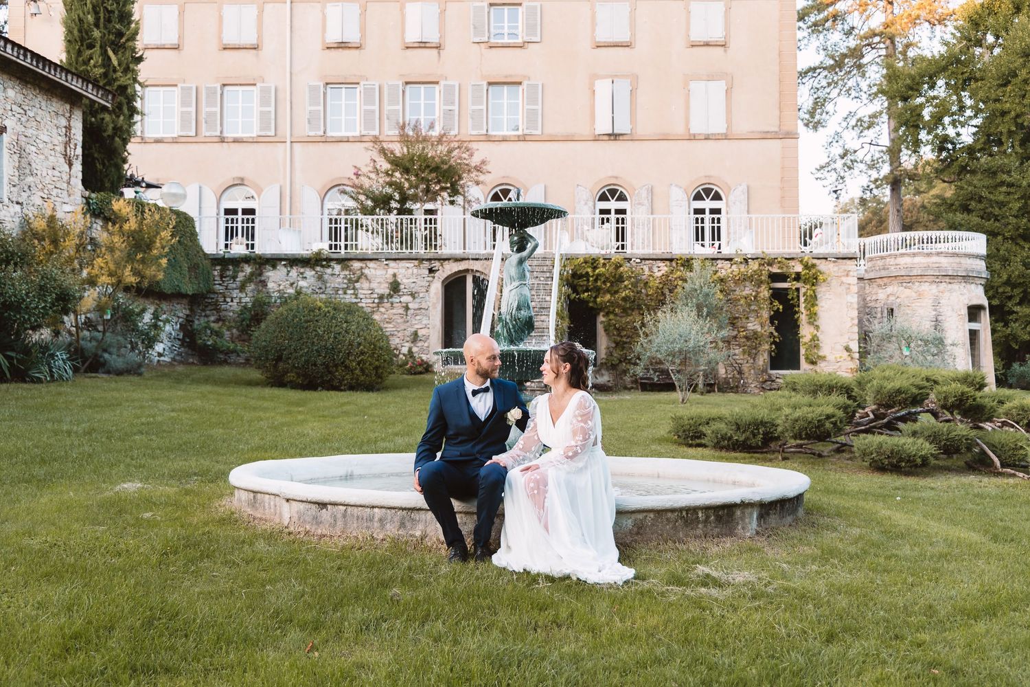 Wedding couple seated at vintage stone fountain before a grand French chateau surrounded by lush gardens.