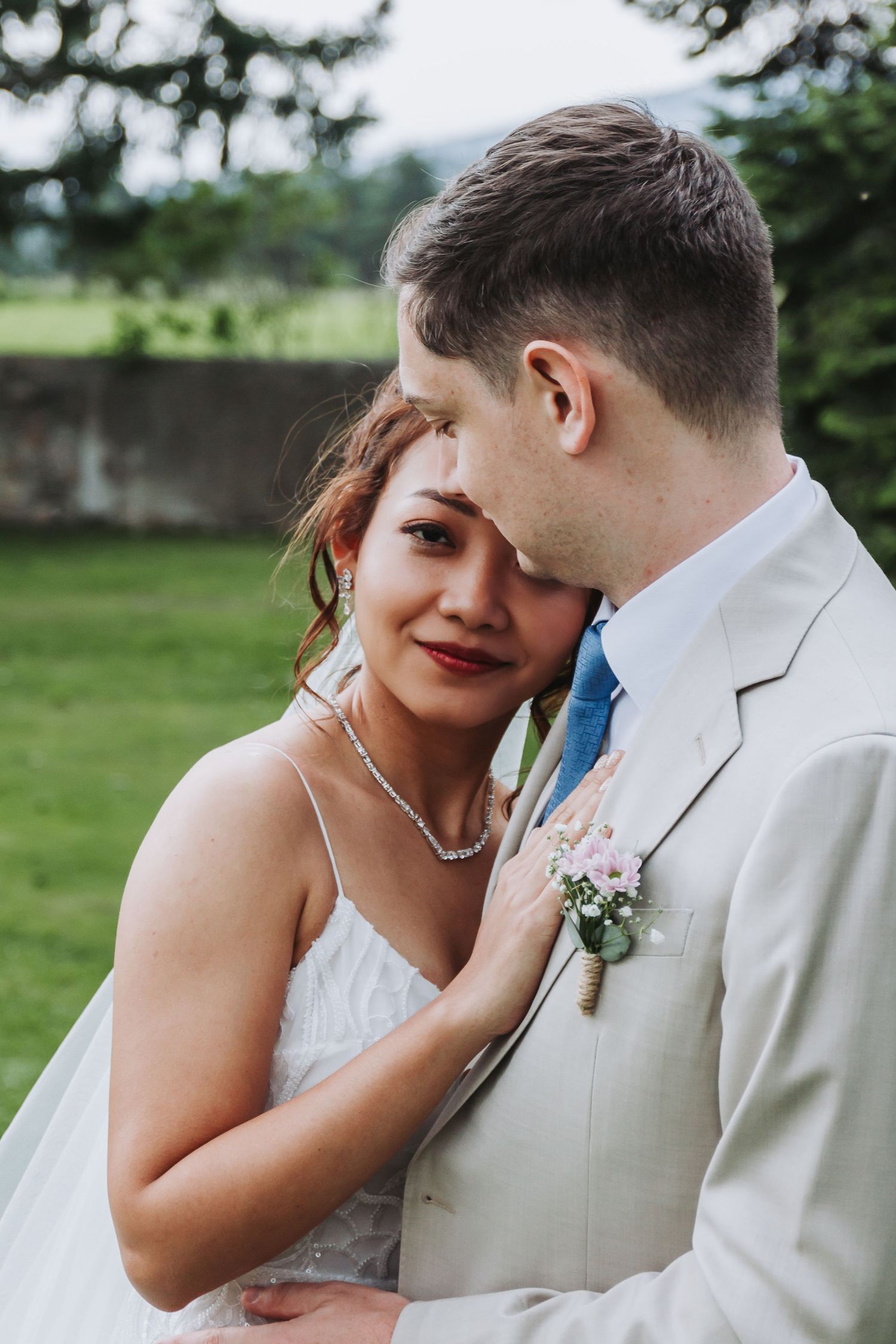Bride in white lace dress leans against groom in beige suit with floral boutonniere during outdoor wedding portrait.