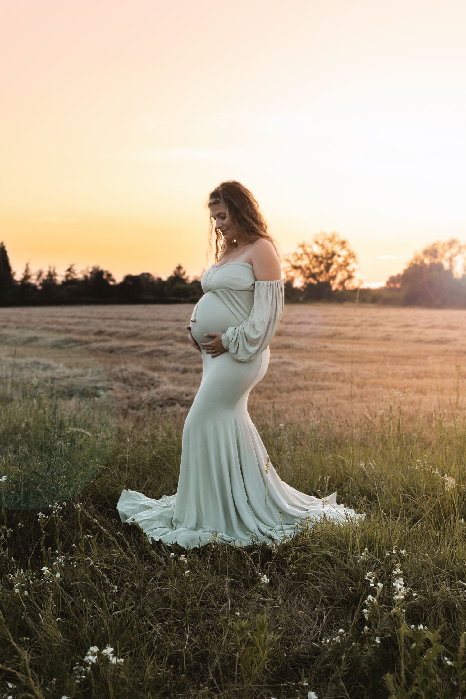 A pregnant woman in a sage green off-shoulder gown poses in a field during a golden sunset.