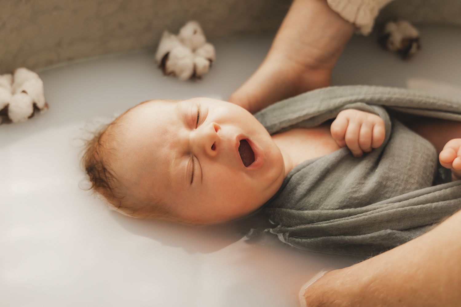 Newborn baby yawning during bath, wrapped in green muslin cloth in milky water.