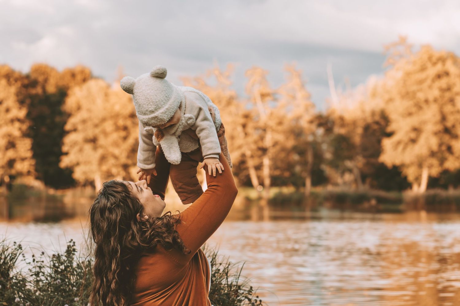 Mother lifting happy baby in the air by an autumn lake with golden foliage background.