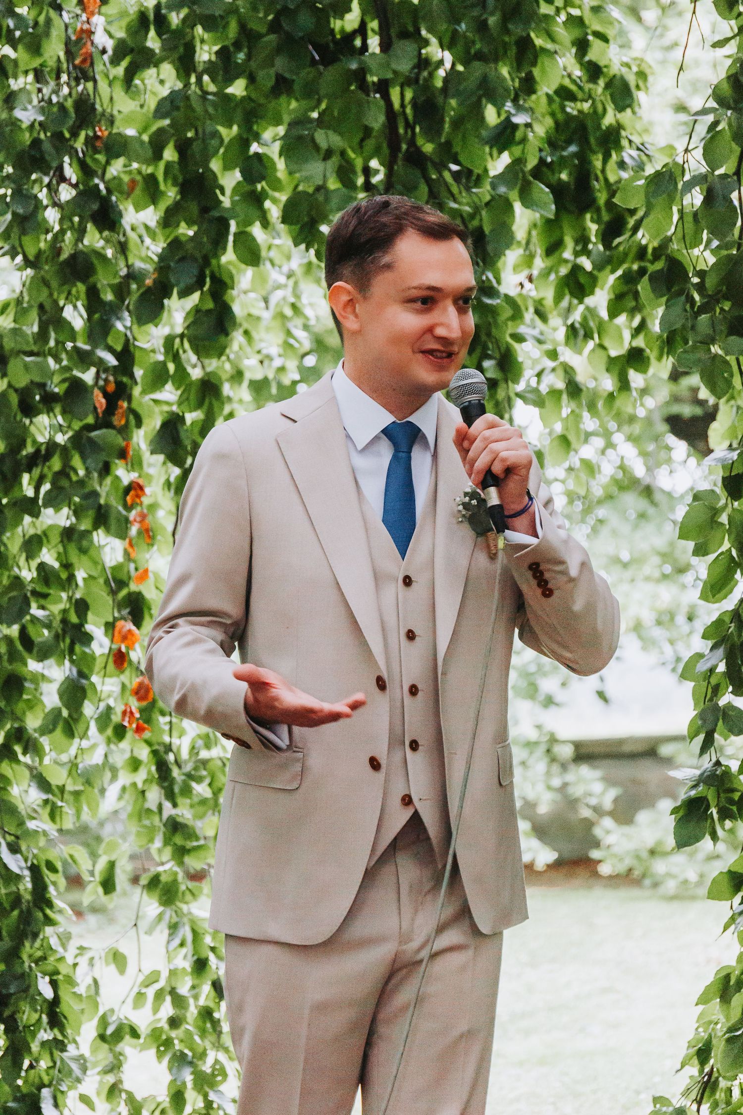 Groom in beige suit and blue tie speaks into microphone during outdoor wedding ceremony under green foliage.