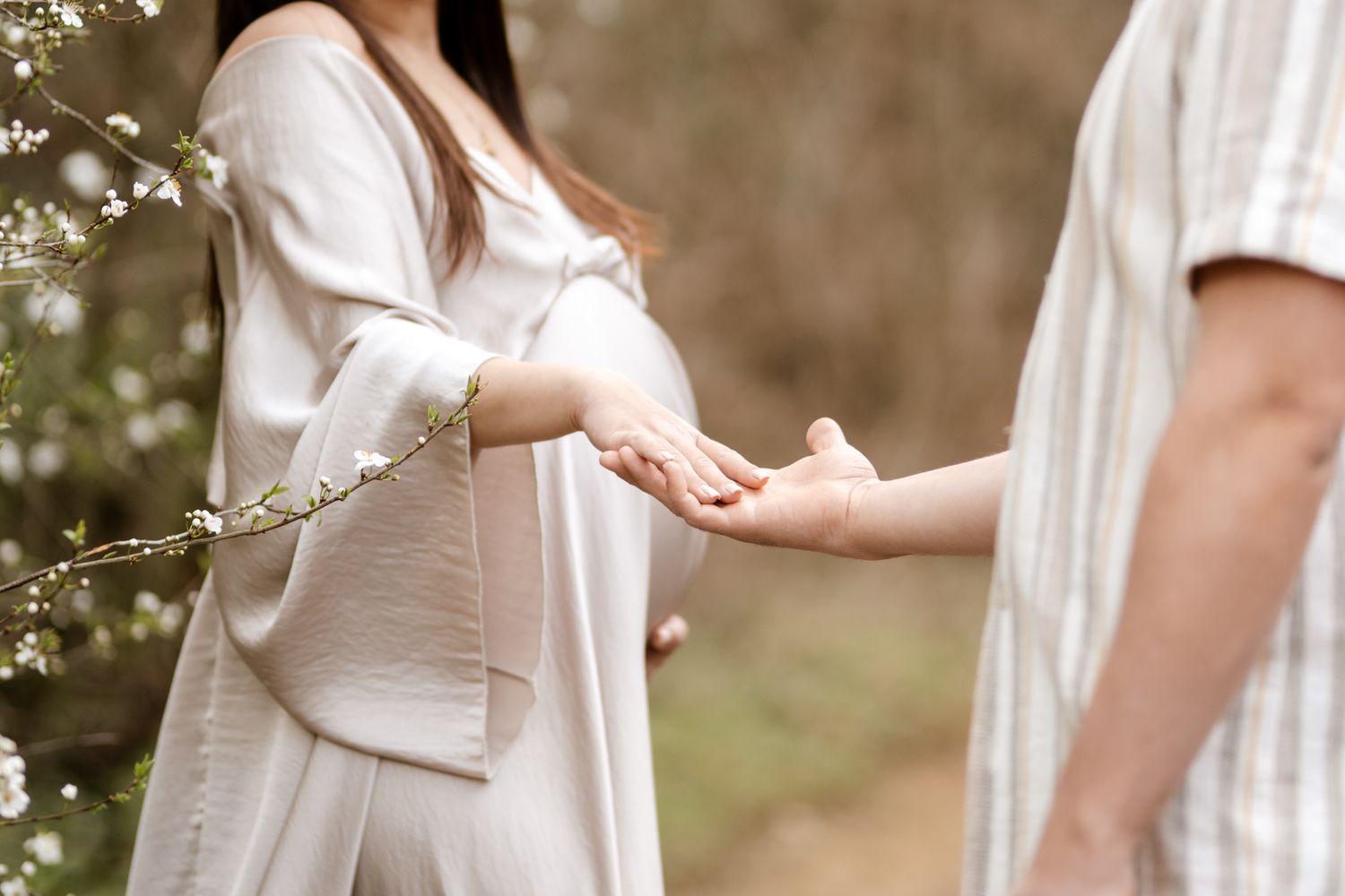 Séance photo grossesse en Isère : un couple se tient la main près d'un arbre en fleurs, future maman en robe nacrée.