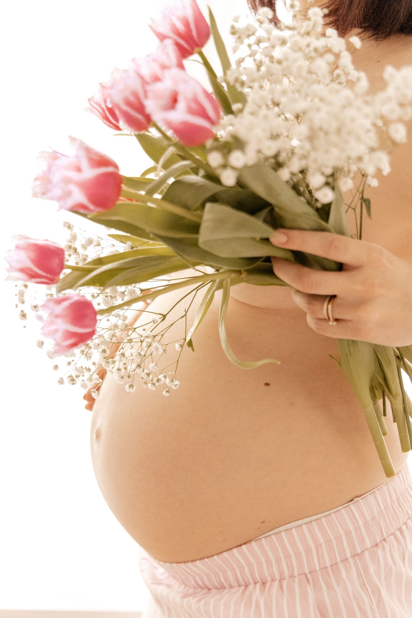 Close-up of pregnant belly with pink tulips and white flowers bouquet in soft light.