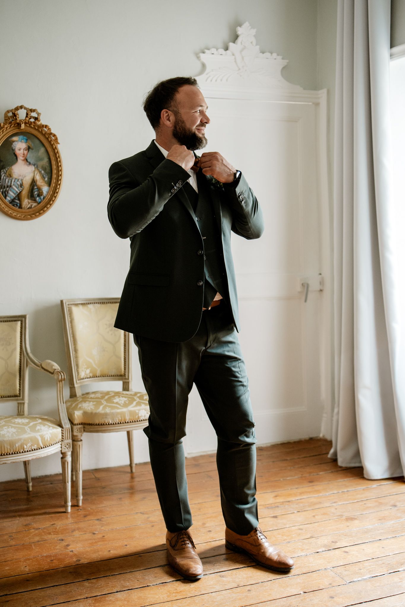 Bearded groom in dark green three-piece suit adjusting his tie in an elegant room with hardwood floors.