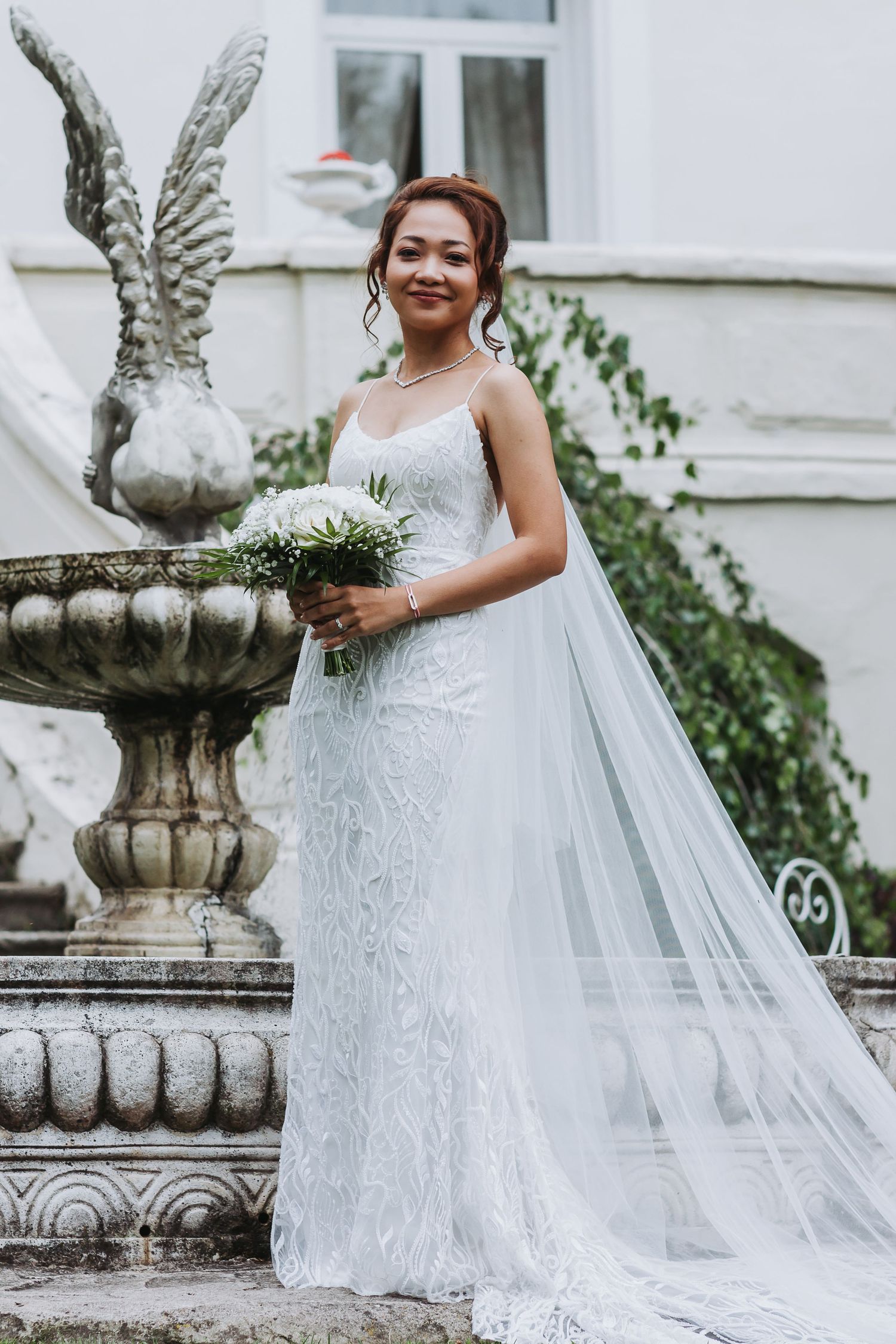 Smiling Asian bride in white lace gown holding bouquet beside ornate stone fountain at elegant venue.
