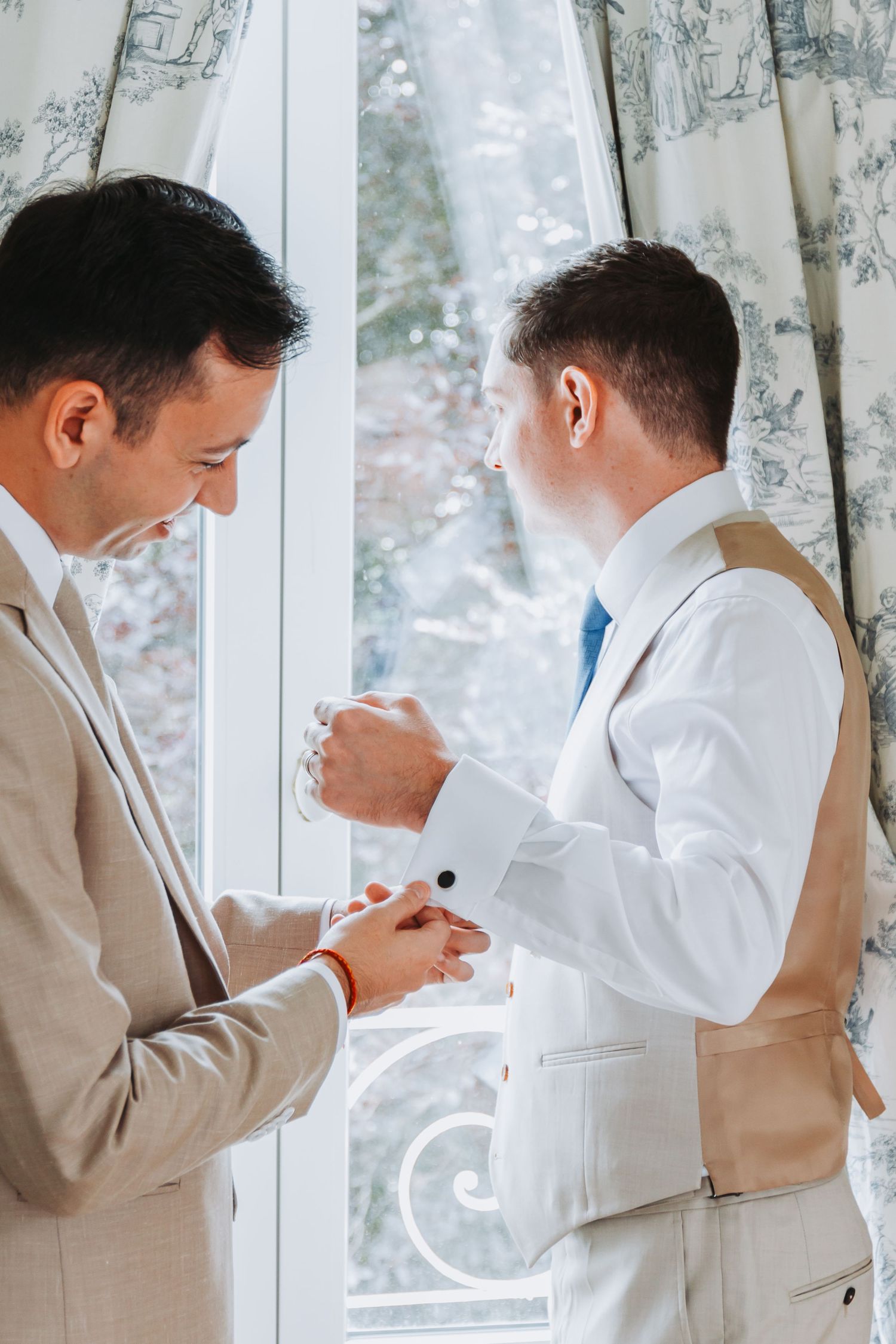 Groomsman helping groom fasten cufflinks while getting ready near a bright window before the wedding.